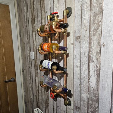 Wall-mounted steampunk wine rack made from copper pipes holding four bottles against rustic wood paneling