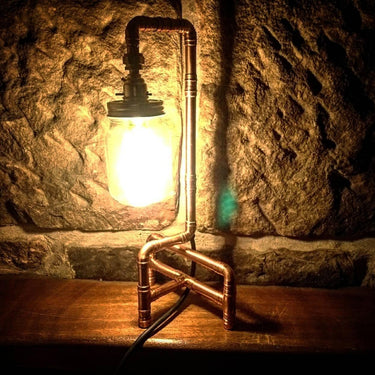 Industrial pipe table lamp with glowing mason jar, set on wood against stone wall