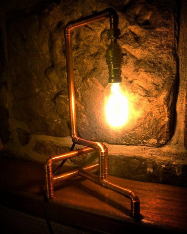 Industrial pipe style table lamp with exposed bulb on a wooden shelf against a stone wall
