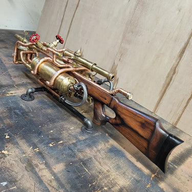 Steampunk-style brass and copper pipe rifle prop with a dark wood stock, mounted on a black support stand against a beige wall.