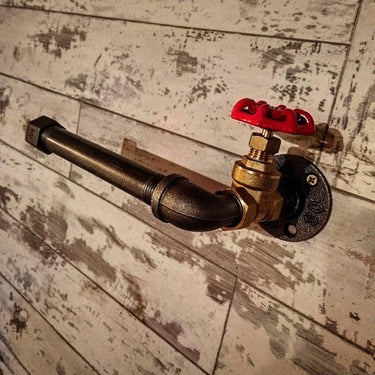 Steampunk industrial pipe toilet paper holder with red valve on rustic wood wall.