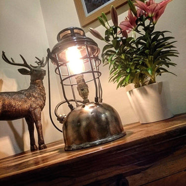Vintage industrial table lamp with exposed bulb on wooden table, next to metal deer figurine and potted flowers.