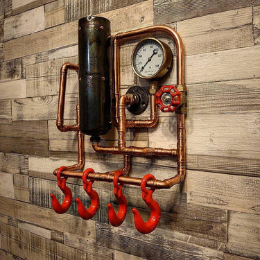 Industrial steampunk coat rack with copper pipes, pressure gauge, and red hooks on wood wall.