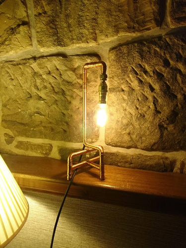 Industrial pipe style table lamp with exposed bulb on wooden shelf against stone wall
