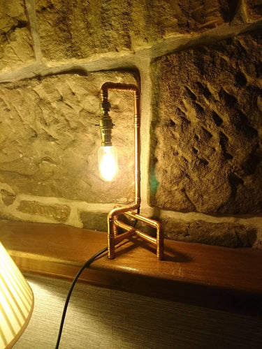 Industrial pipe style table lamp with exposed bulb on wooden shelf against stone wall