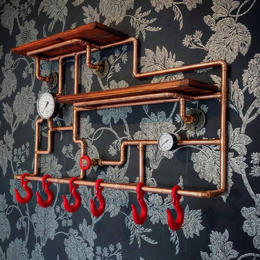 Steampunk coat rack with copper pipes, red hooks, wooden shelves, and vintage gauges on floral wallpaper.