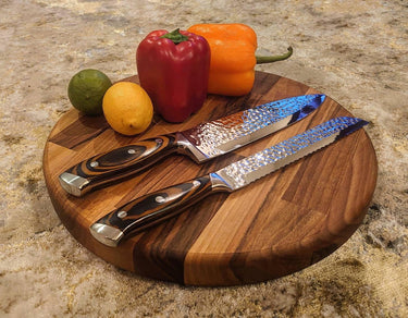 Round chunky walnut chopping board with two knives, vegetables, and citrus fruits on top