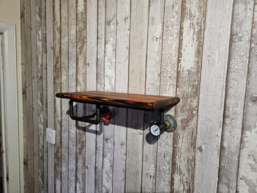 Handmade industrial wall shelf with wood plank and black metal pipe fittings, pressure gauge detail, mounted on rustic wood panel wall.