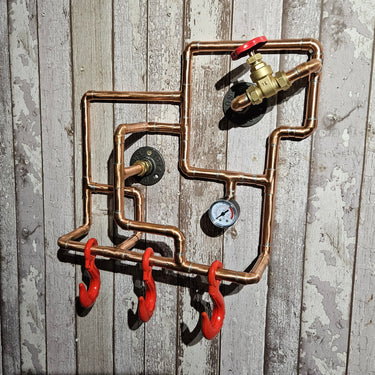 Steampunk copper pipe wall rack with red hooks, industrial gauge, and valve on rustic wood panel