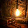 Industrial pipe style table lamp with exposed bulb on a wooden shelf against a stone wall