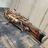 Steampunk-style brass and copper pipe rifle prop with a dark wood stock, mounted on a black support stand against a beige wall.