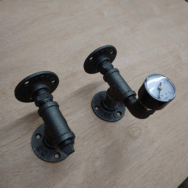 Pair of black steampunk industrial pipe wall brackets, one with a vintage gauge, on wood surface