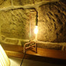 Industrial pipe style table lamp with exposed bulb on wooden shelf against stone wall