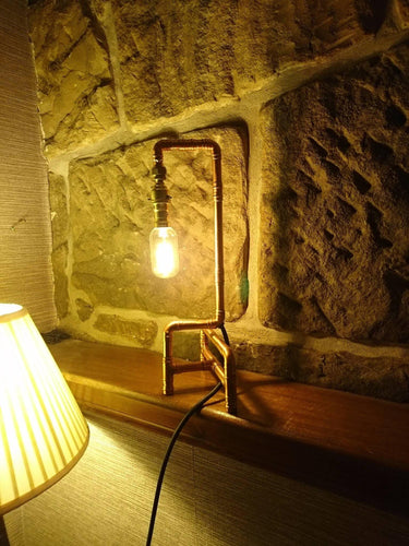 industrial pipe table lamp with exposed bulb on wooden shelf against stone wall