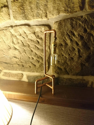 Tall industrial pipe table lamp with exposed Edison bulb on a wooden shelf against stone wall