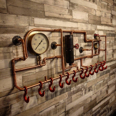 Steampunk coat rack made from copper pipes, pressure gauges, and red hooks mounted on wood wall.
