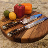 Round chunky walnut chopping board with two knives, vegetables, and citrus fruits on top